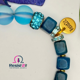 Close-up of a blue beaded bracelet with decorative elements and a Resist2it charm, featuring theResist2it brand.
