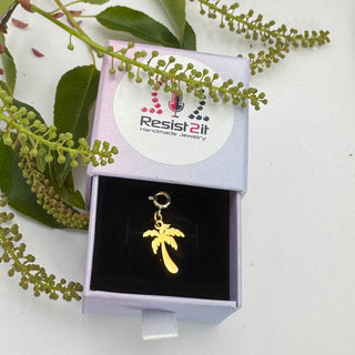 Gold palm tree charm in a box with 'Resist2it' branding, surrounded by green leaves.