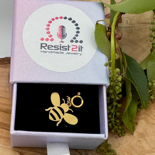 Jules' Golden Bee™ charm in a box with 'Resist2it Handmade Jewelry' logo, surrounded by green leaves.