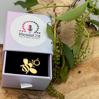 Jules' Golden Bee™ charm in a 'Resist 2it' jewelry box on a wooden surface with green leaves.