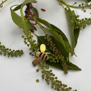Jules' Golden Bee™ charm on a branch with green leaves and small flowers on a white background