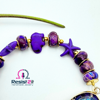 Close-up of part of PURPLE TIDES™ handmade bracelet, showing starfish and elephant and emperor stones with golden seed beads and rondelles with Resist2it logo