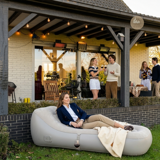 Woman relaxing on a beige inflatable lounger in a garden setting with a house and outdoor gathering — Sun Drift Lounger™
